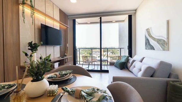 Un comedor moderno con una mesa para dos, un sofá, un televisor en la pared y un balcón con vistas.
