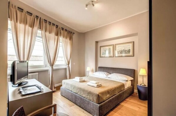 A bedroom with a bed, a desk with a computer, a lamp, and a window with curtains. - The Dome (Roma)