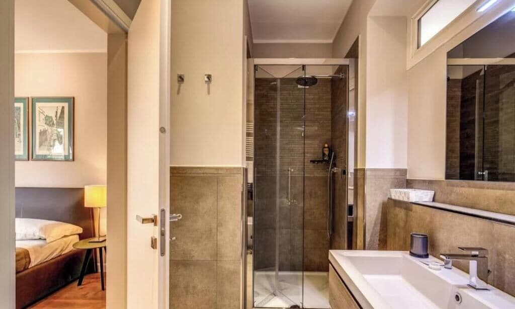 Habitación de hotel con ducha envuelta en vidrio, un espejo grande y una cama con mesita de noche.