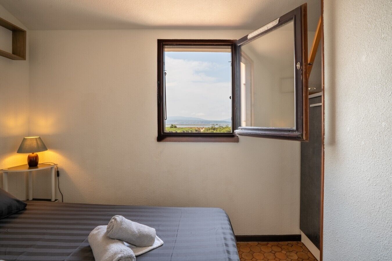 Una camera d'hotel con un letto, un comodino con una lampada, una finestra vista sul paesaggio e una porta.