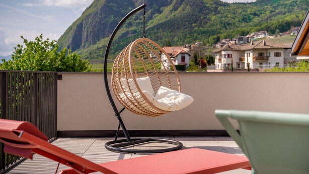 Uma cadeira suspensa de vime com almofadas em uma varanda com vista para uma paisagem montanhosa.