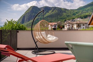 Ein hängender Korbstuhl mit Kissen auf einem Balkon mit Blick auf eine gebirgige Landschaft.