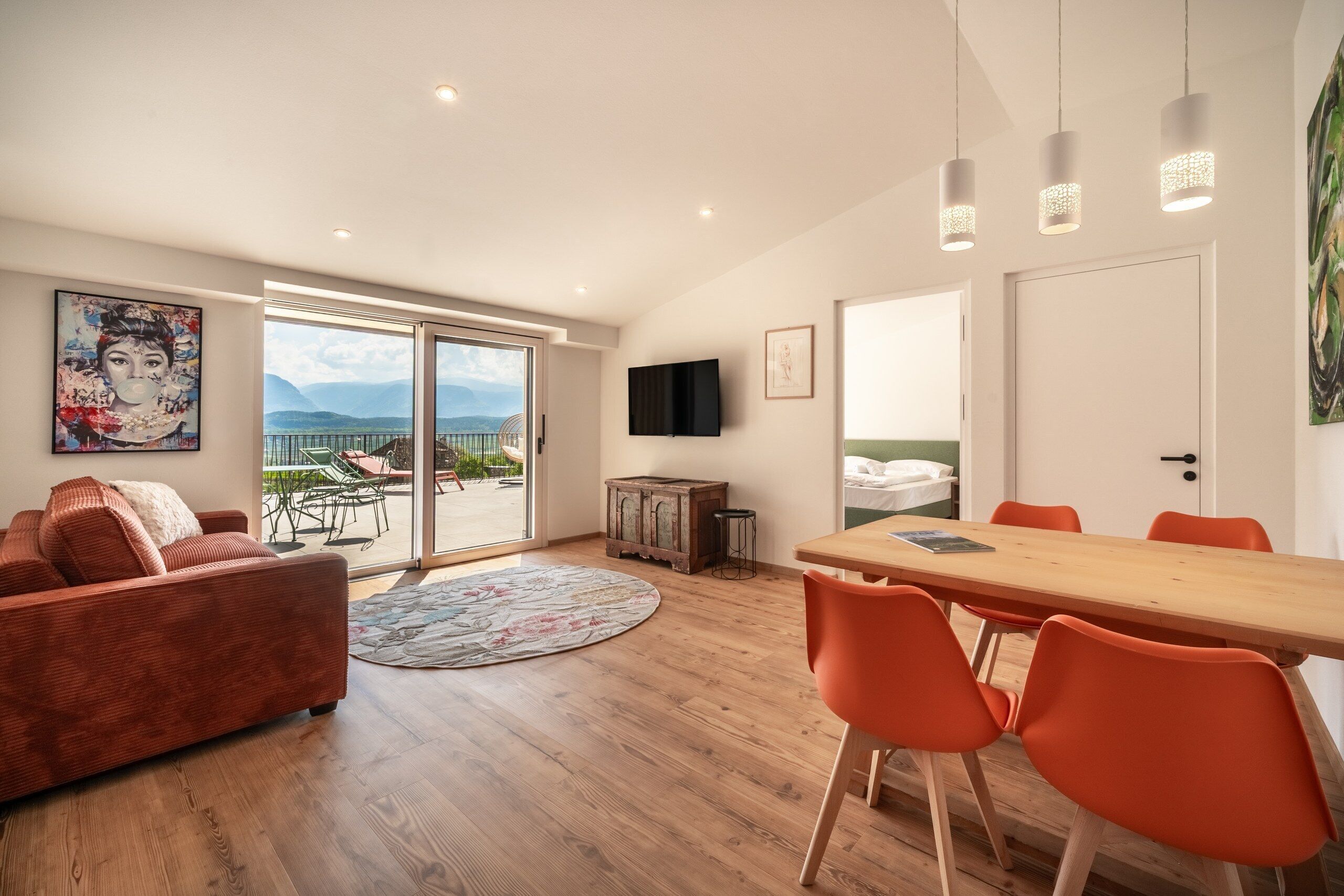 Ein modernes Wohnzimmer mit großem Fenster, einem Essbereich mit orangefarbenen Stühlen und einem an der Wand montierten Flachbildfernseher.