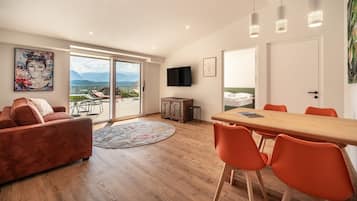 Ein modernes Wohnzimmer mit großem Fenster, einem Essbereich mit orangefarbenen Stühlen und einem an der Wand montierten Flachbildfernseher.
