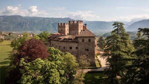Um castelo de pedra histórico com ameias, cercado por vegetação exuberante e montanhas ao fundo.