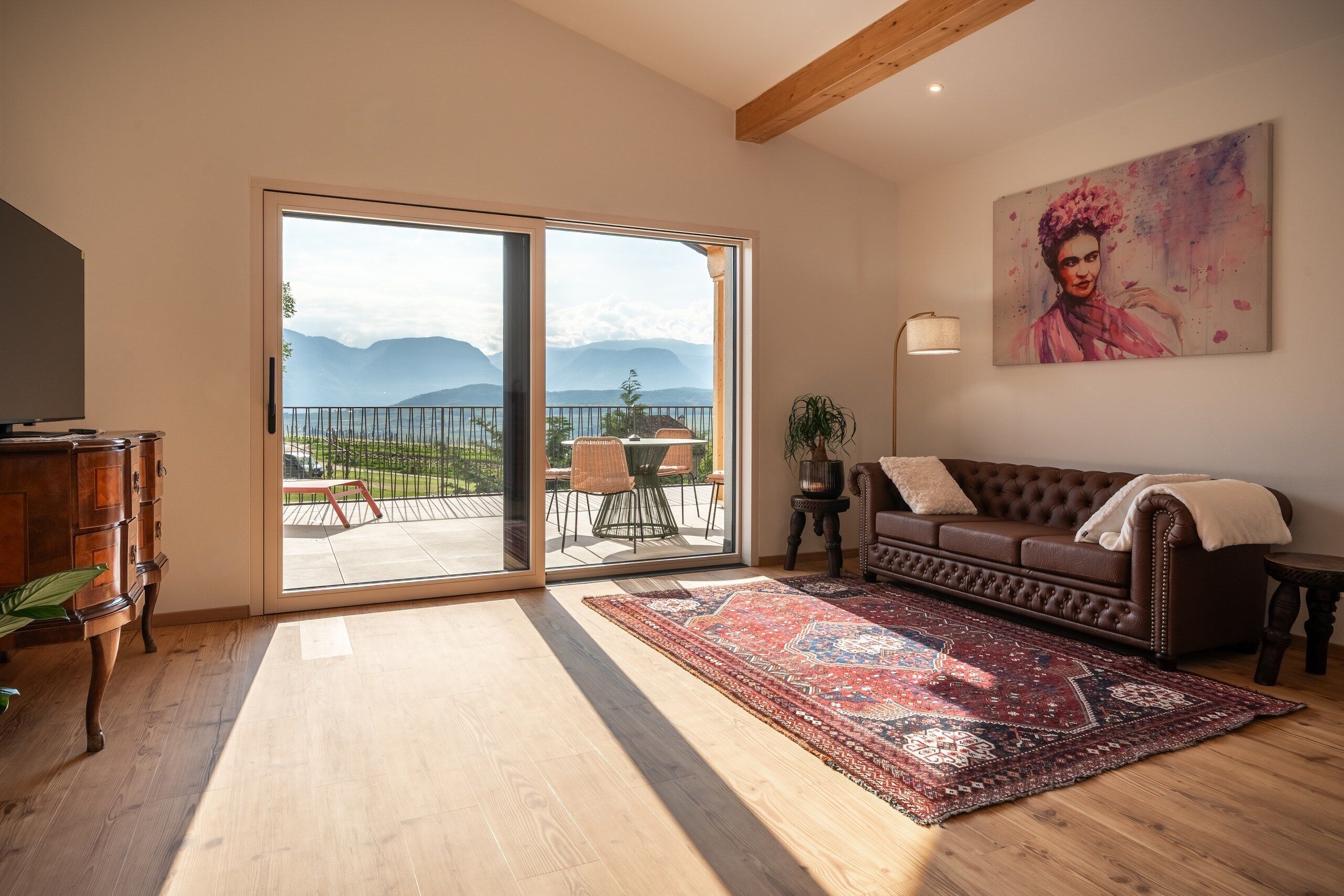Un soggiorno spazioso con un'ampia porta scorrevole in vetro che dà su un balcone, un divano in pelle marrone, un tappeto decorato e un quadro appeso al muro.