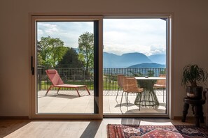 Ein Zimmer mit einer Schiebetür, die auf eine Terrasse mit Tisch und Stühlen, einem roten Liegestuhl und einem Topfpflanzen führt.