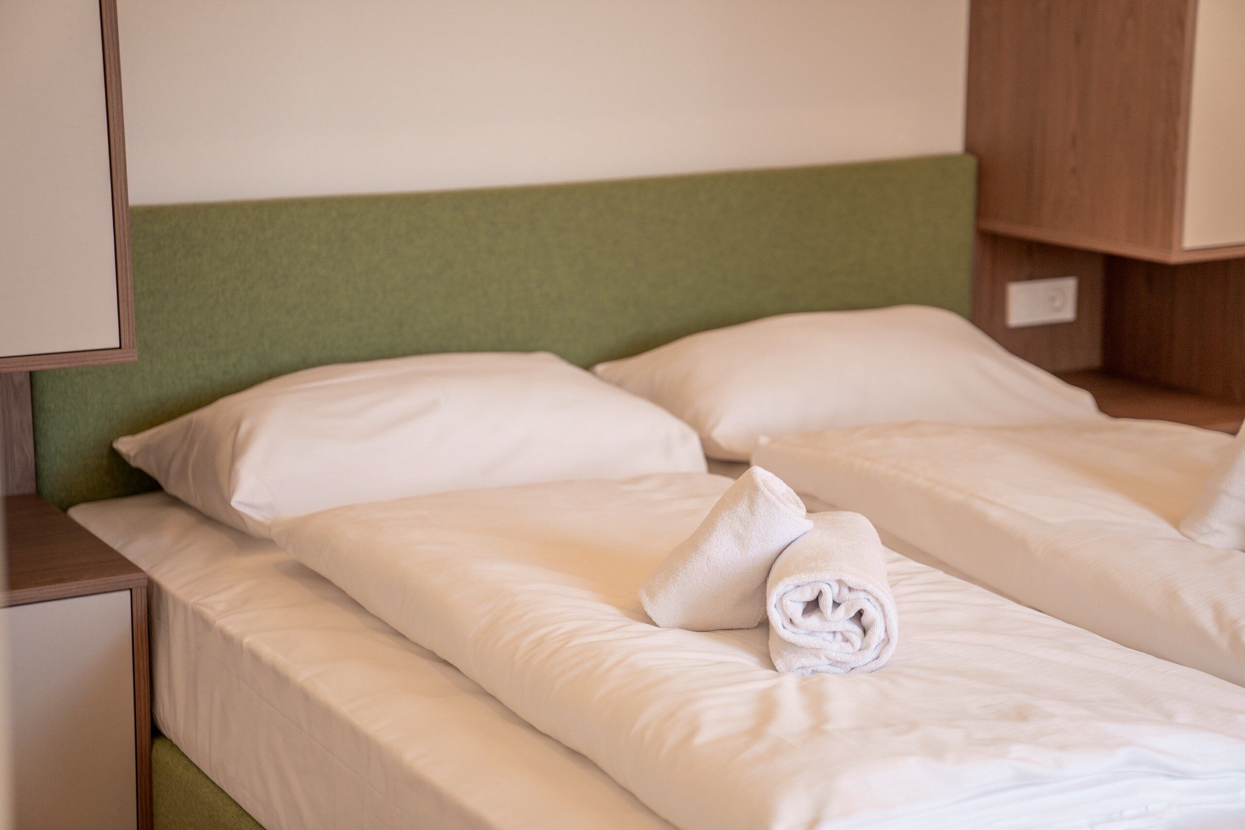 Ein Hotelzimmer mit einem Einzelbett, einem grünen Kopfteil und einem ordentlich bezogenen Bett mit weißen Bettwäsche und einem zusammengerollten Handtuch.