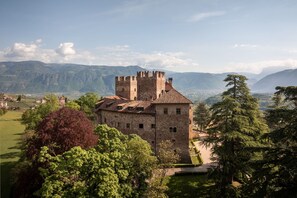 Un château de pierre historique, doté d’une tour, entouré d’une végétation luxuriante et avec des montagnes en toile de fond.