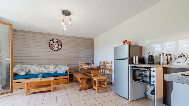 A compact living space with a sofa, a dining table, chairs, a kitchen area with a refrigerator and oven, and a wall clock.