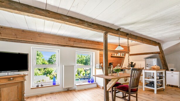 Un loft espacioso con techo alto, vigas de madera, zona de comedor con mesa y sillas, cocina con armarios blancos y ventanas amplias con vistas al verdor.