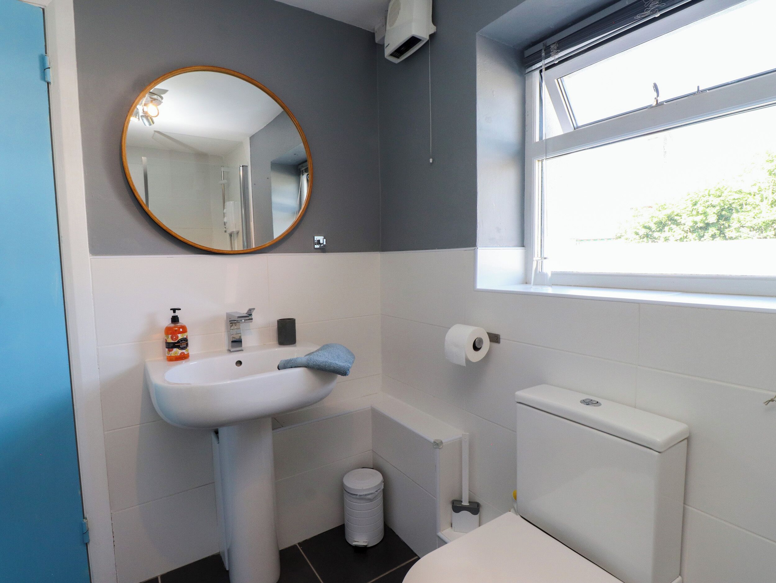 A bathroom with a pedestal sink, a toilet, a bidet, a trash bin, a mirror, and a window.