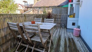 En träveranda med ett bord och stolar, en rosa tunna och en byggnad med ett grönt tak i bakgrunden.