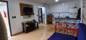 A living room with a TV, a sofa, a kitchen area with a tiled backsplash, and a wooden cabinet. - Apartment for 6 people in the center of Bombinhas with breakfast. (Bombinhas)