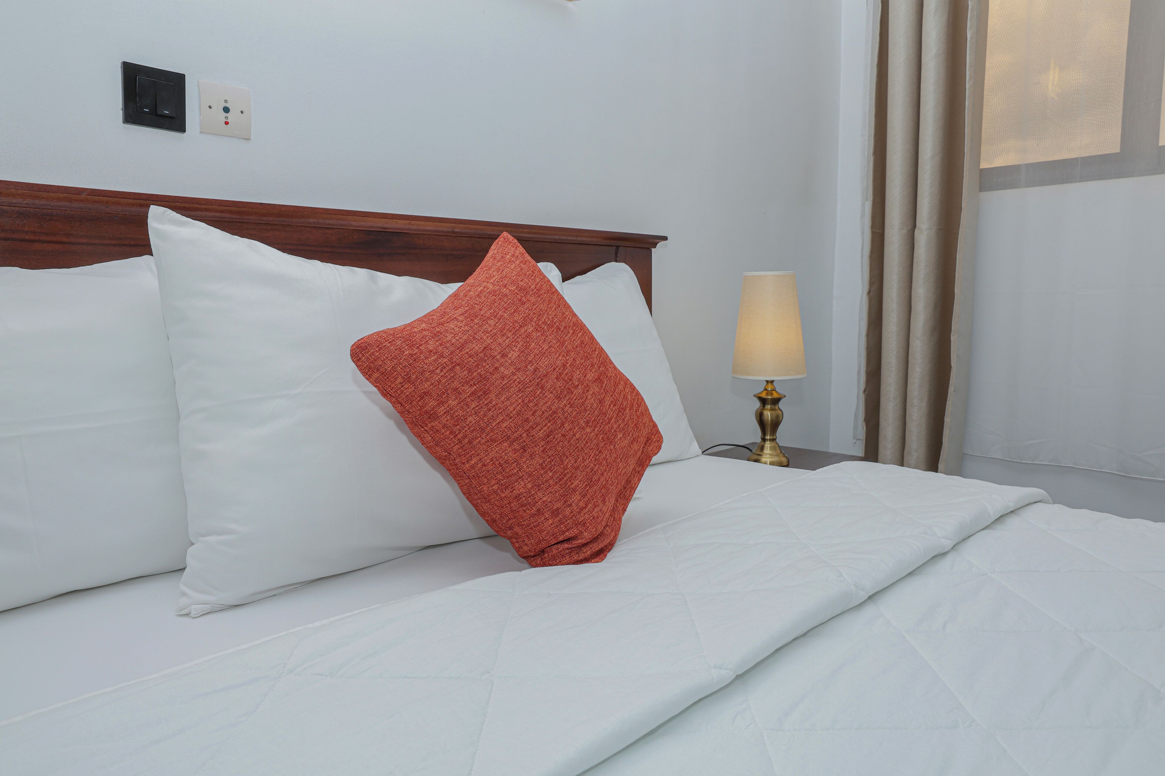 Habitación de hotel con cama, ropa de cama blanca, almohada roja, lámpara de noche y cabecera de madera.