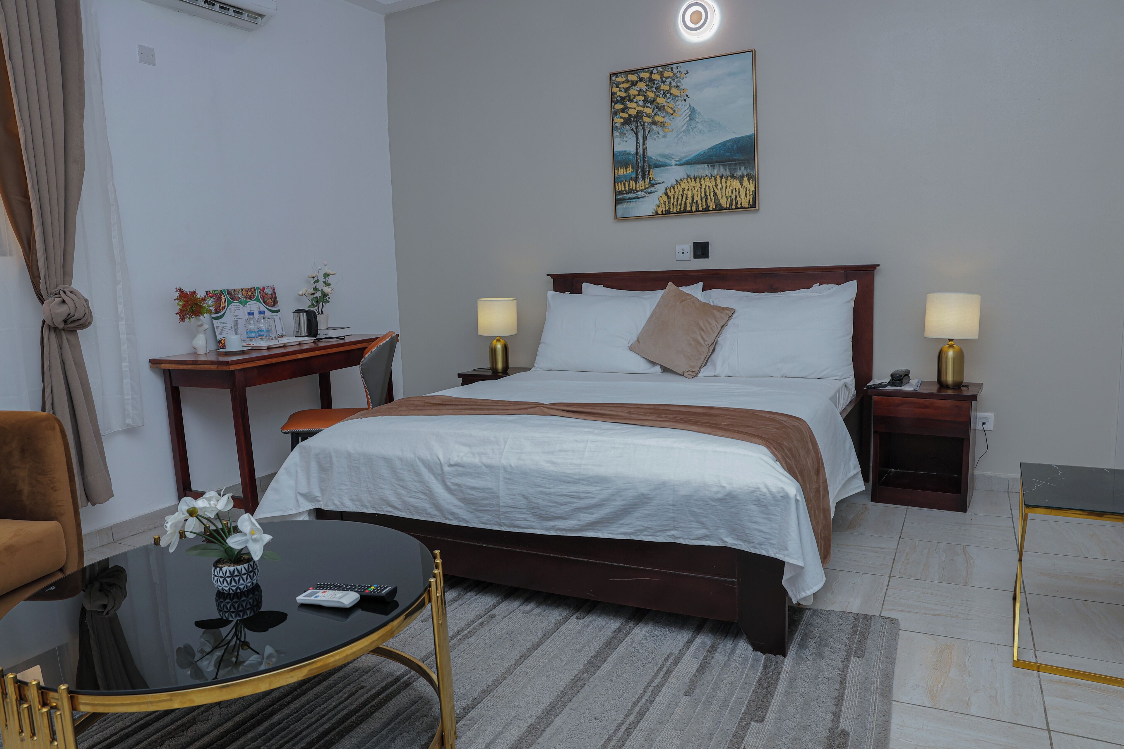 A hotel room with a bed, bedside tables, a desk, a chair, and a painting on the wall.