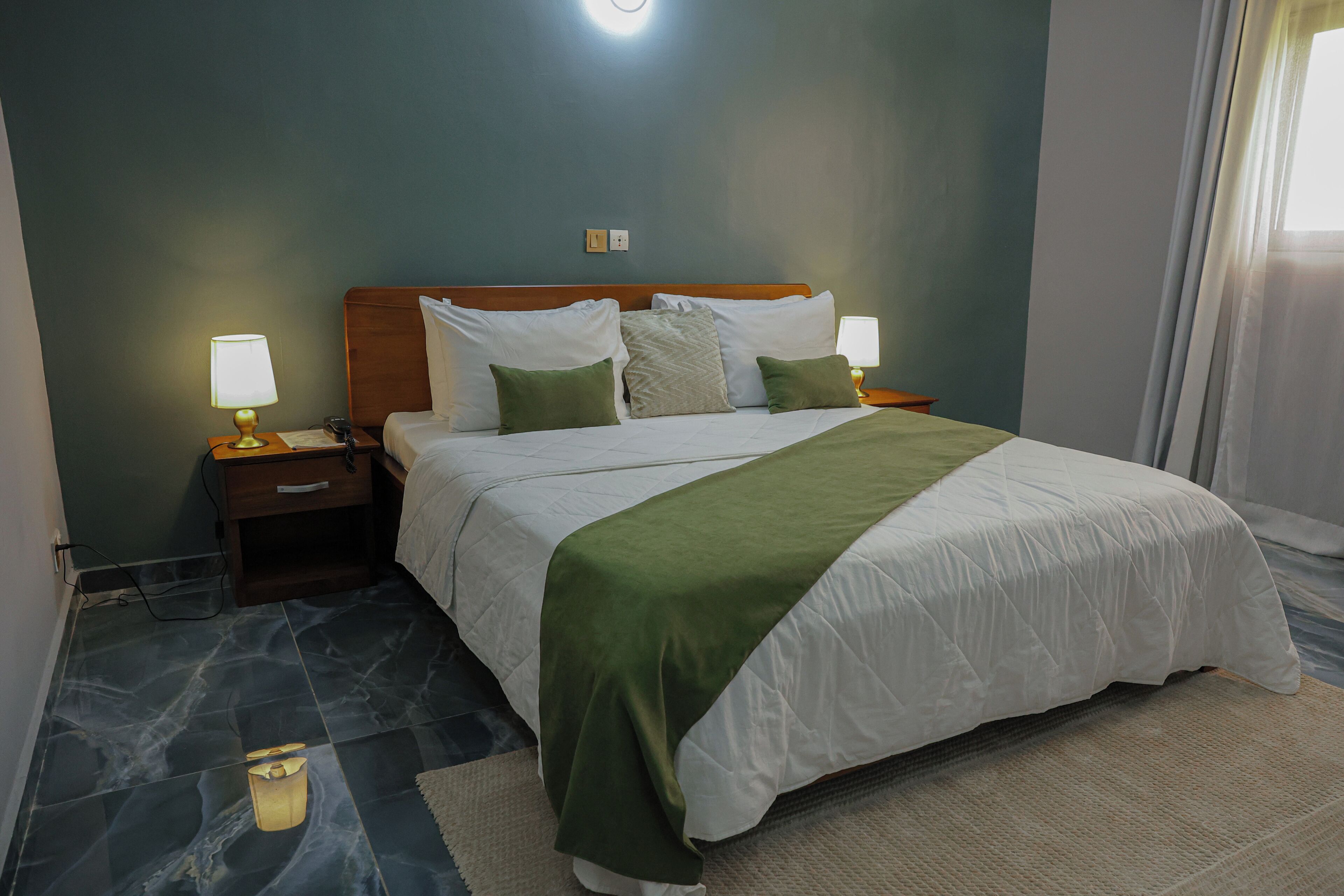 A hotel room with a bed, two bedside tables with lamps, a nightstand, a wall socket, and a window with curtains.