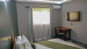 A hotel room with a bed, a desk, a chair, a TV, and an air conditioning unit. - Les jardins d'Adzopé (Adzope)