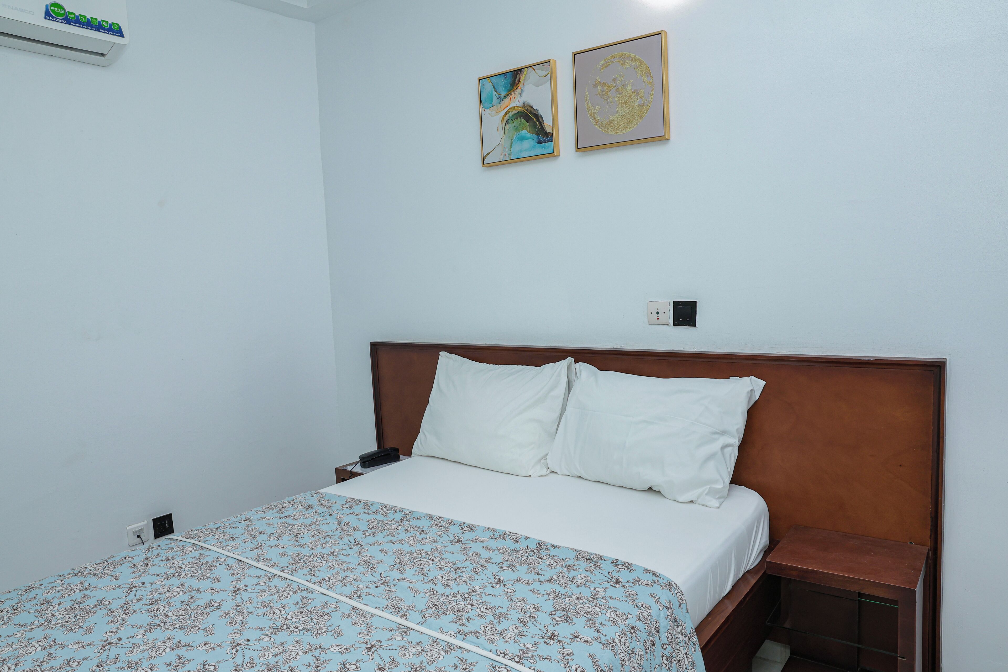 Habitación de hotel con cama, mesita de noche, aire acondicionado instalado en la pared y dos obras de arte enmarcadas.