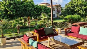 A terrace with wicker furniture, a glass table, and a view of a house and greenery.
