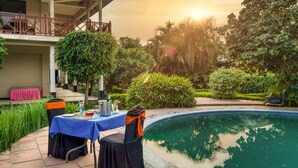 A poolside area with a table and chairs, a covered seating area, and lush greenery.