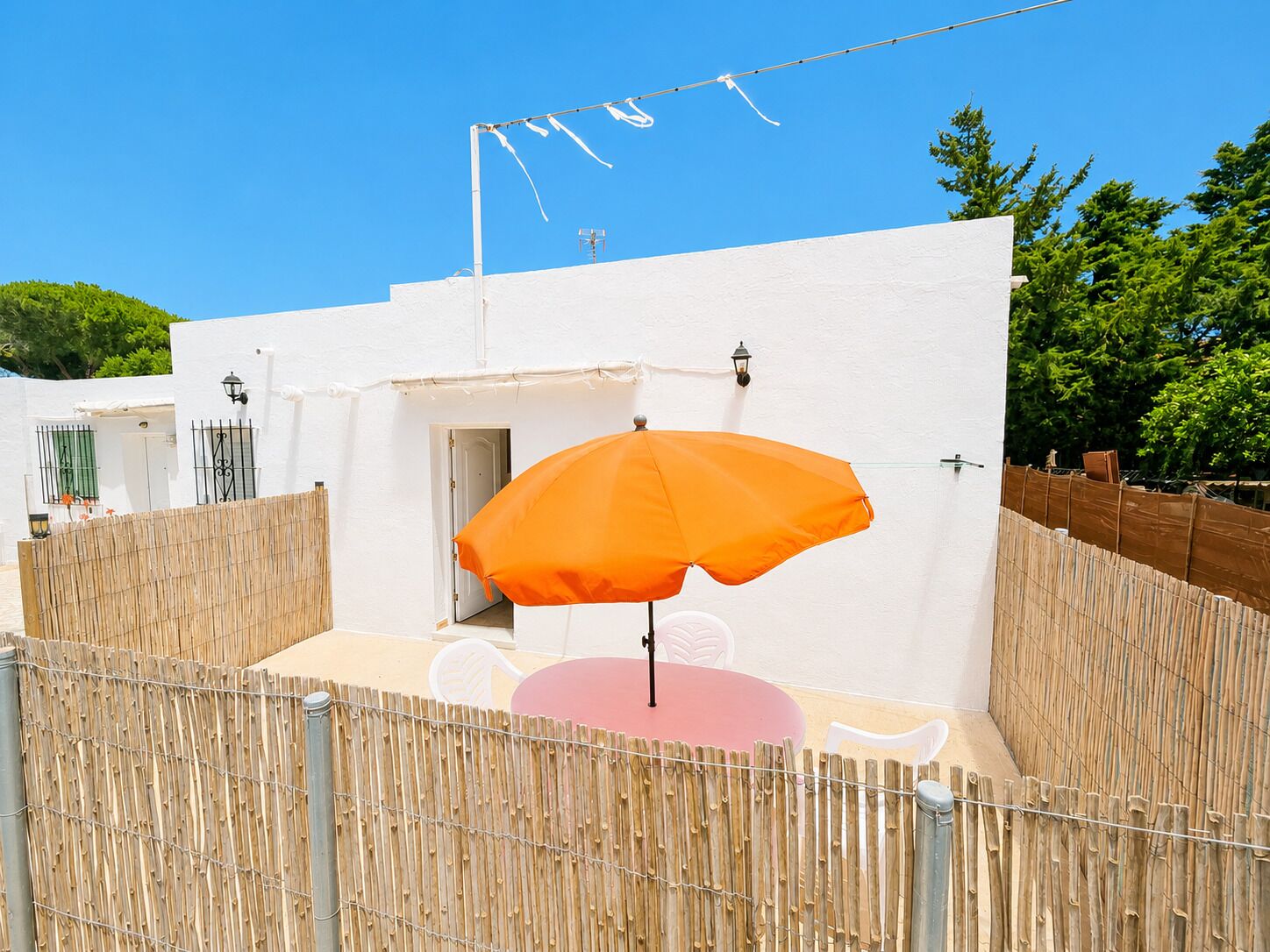 Un edificio blanco con valla de bambú, una sombrilla naranja y una mesa rosa.