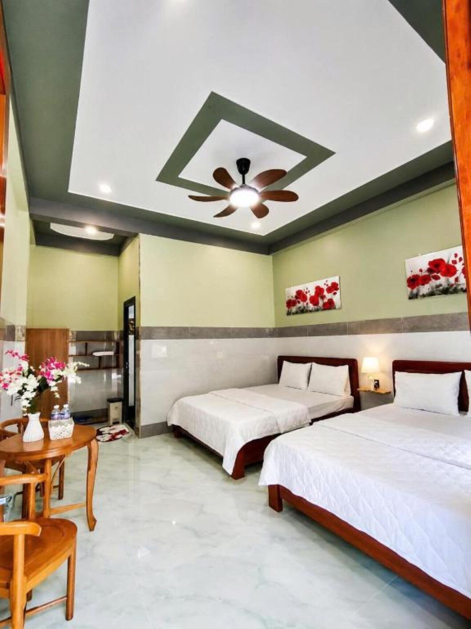 A hotel room with two beds, a ceiling fan, a dining table with chairs, and artwork on the walls.