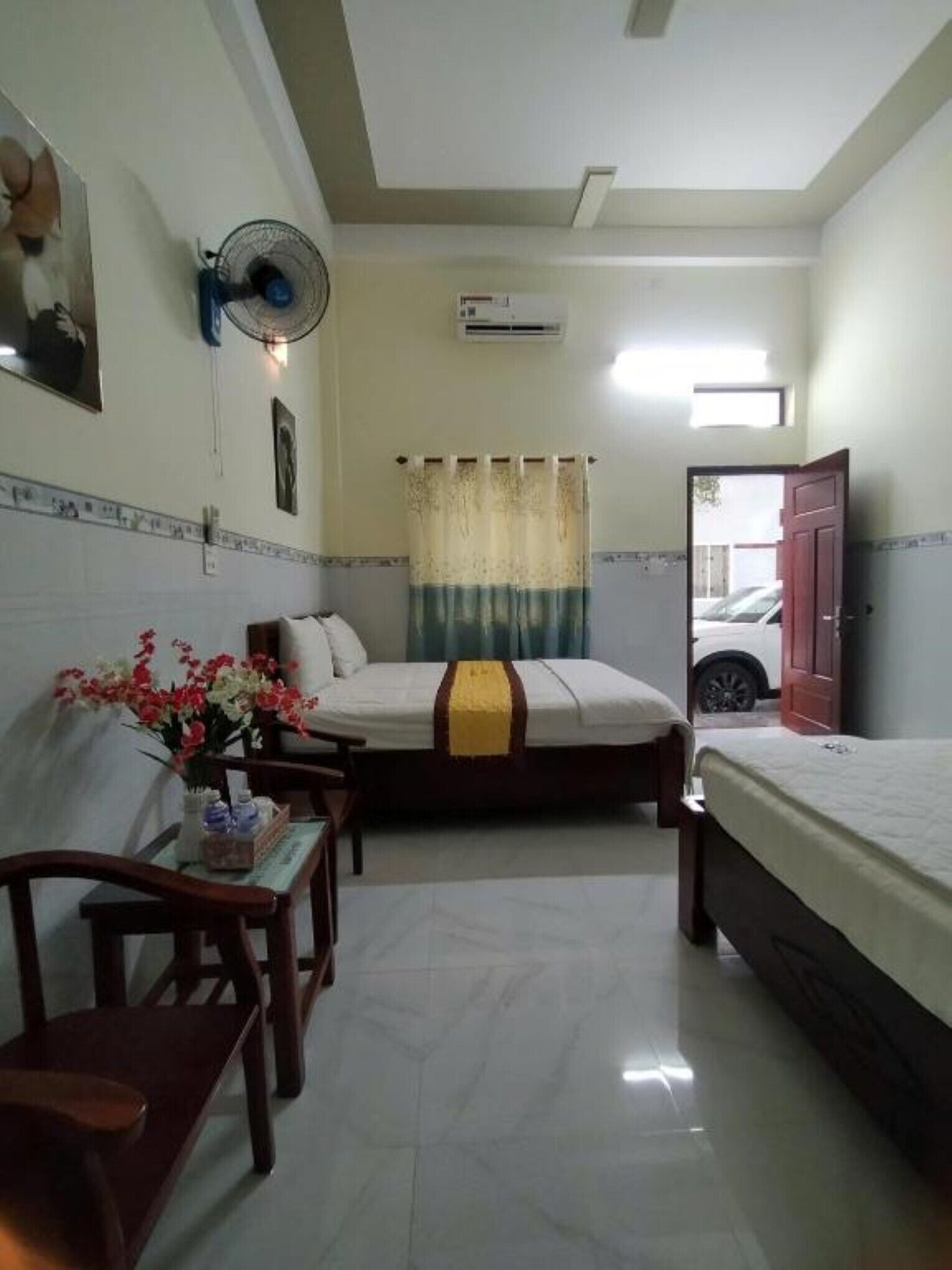 A hotel room with a bed, a desk with a chair, a small table with flowers, a fan, and a window with curtains.