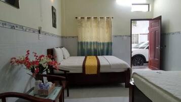 A hotel room with a bed, a desk with a chair, a small table with flowers, a fan, and a window with curtains.
