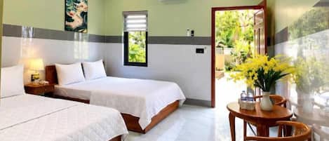 A hotel room with two beds, a ceiling fan, a window with blinds, a table with chairs, and a view of greenery outside.