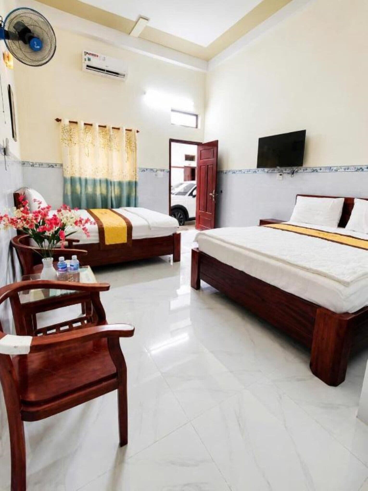 A hotel room with two beds, a wooden chair, a table with a vase of flowers, and an air conditioning unit.
