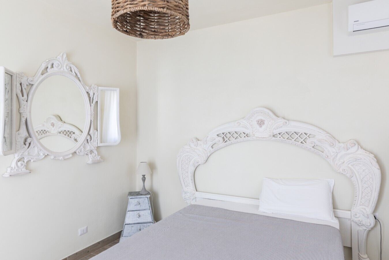 A bedroom with a white ornate headboard, a white bedspread, a white dresser, a white lamp, and a white mirror with an ornate frame.