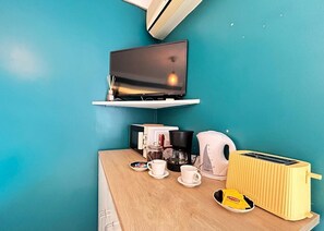 Una habitación con pared color turquesa, un televisor de pantalla plana sobre una estantería blanca, una cafetera, un hervidor, un tostador y una caja de té Lipton sobre una mesa de madera.