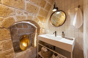 A bathroom with a stone wall, a white sink, a mirror, a towel rack, and a shelf with baskets. - Casa Volta Nova – Historic Hideaway (Polignano a Mare)
