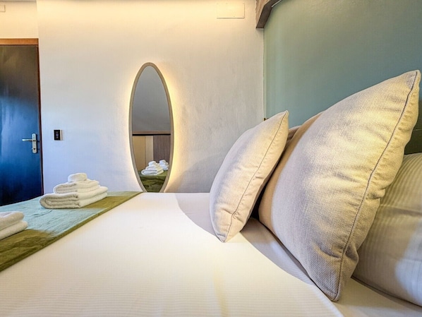 A hotel room with a bed, pillows, a wooden side table with towels, and a mirror with an arch design.