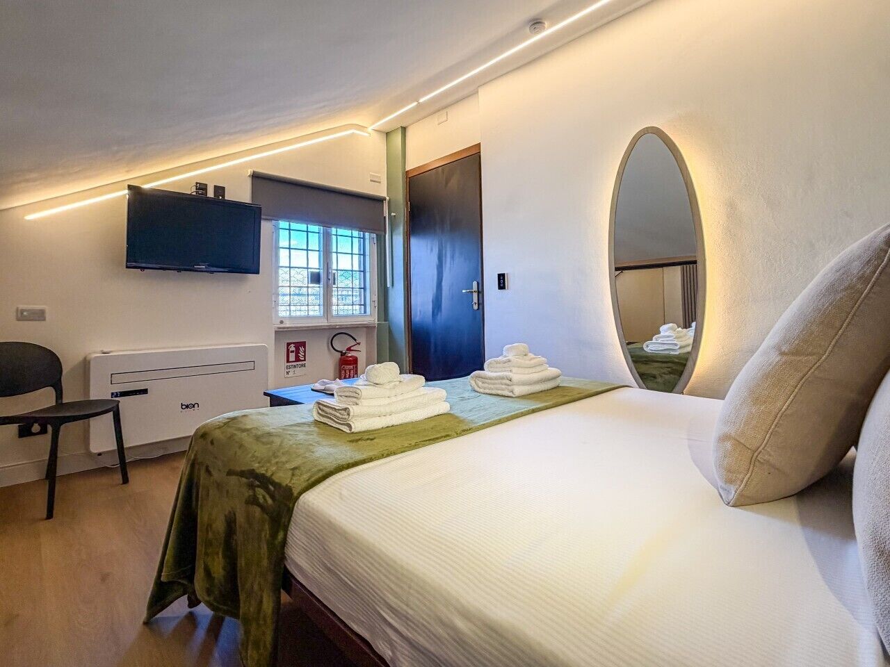 Una camera d'albergo con un letto, una sedia, una scrivania, una televisione, uno specchio e una finestra con vista.