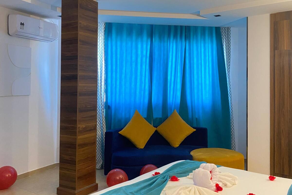 A hotel room with a bed, a blue curtain, a wooden pillar, and a wall-mounted air conditioner.
