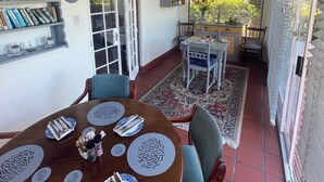 Un patio cubierto con una mesa de comedor para cuatro personas, sillas, una estantería y un ventanal con vistas a la vegetación.