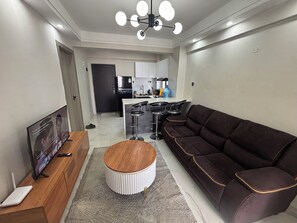 A modern living room with a sofa, a coffee table, and a TV. There is a kitchen area in the background with a fridge and a water dispenser. - Kindaruma Homes, 5 minutes away from yaya centre (Nairobi)