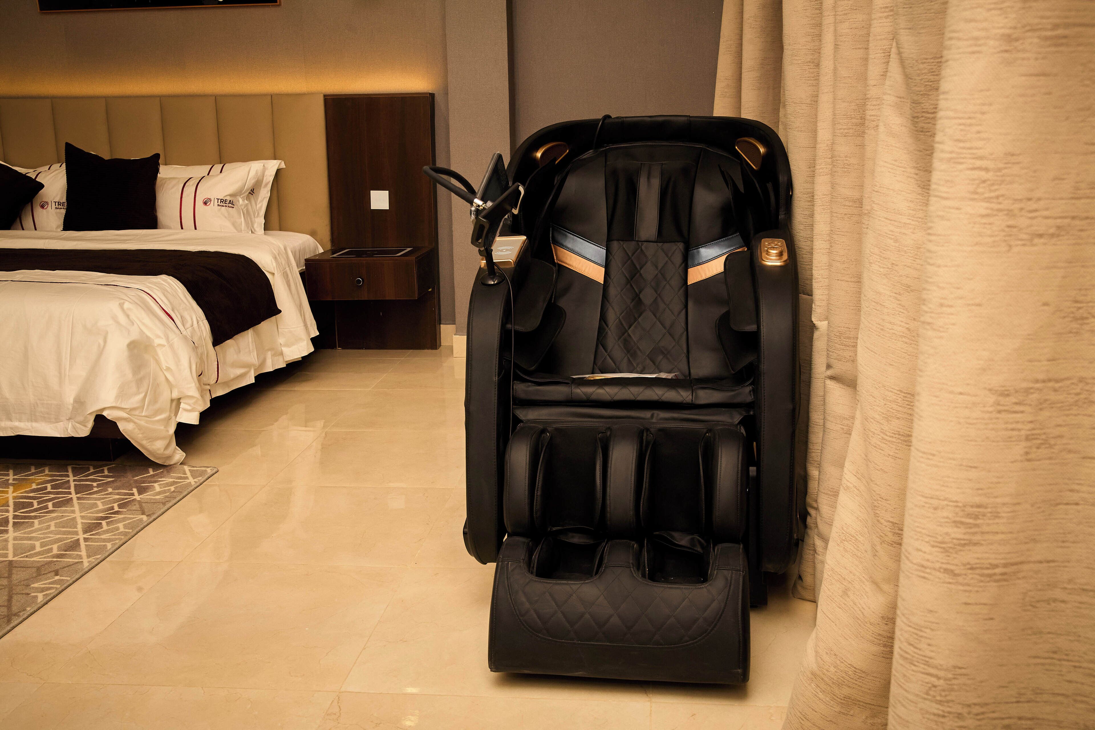Een hotelkamer met een massagestoel, een bed met wit beddengoed, een nachtkastje en een houten hoofdbord.