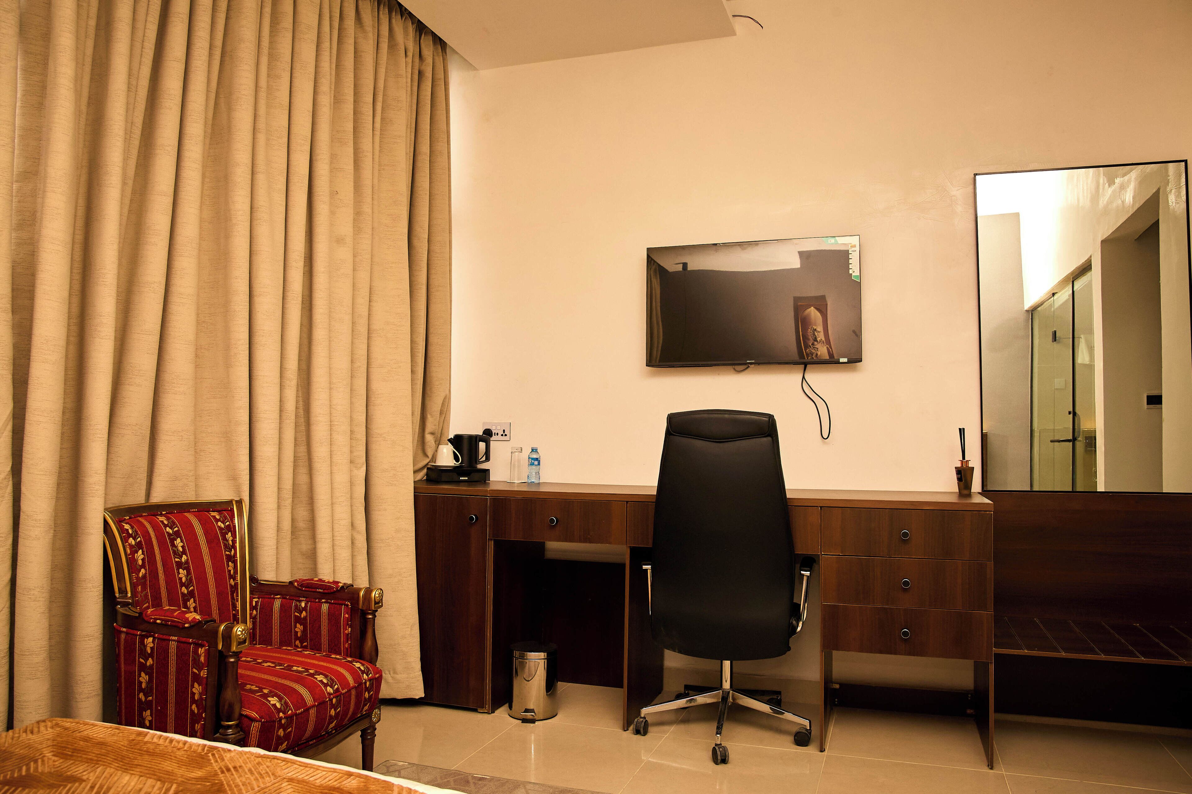 A hotel room with a wooden desk, a black office chair, a television mounted on the wall, a red upholstered chair, and a large mirror.