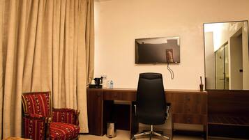 A hotel room with a wooden desk, a black office chair, a television mounted on the wall, a red upholstered chair, and a large mirror.