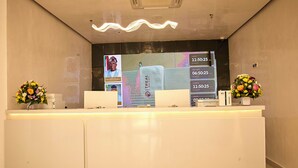 Una recepción con un panel digital, dos arreglos florales y una lámpara moderna.
