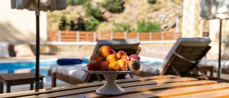 A wooden table with a fruit bowl in the center, a pool in the background, and lounge chairs.