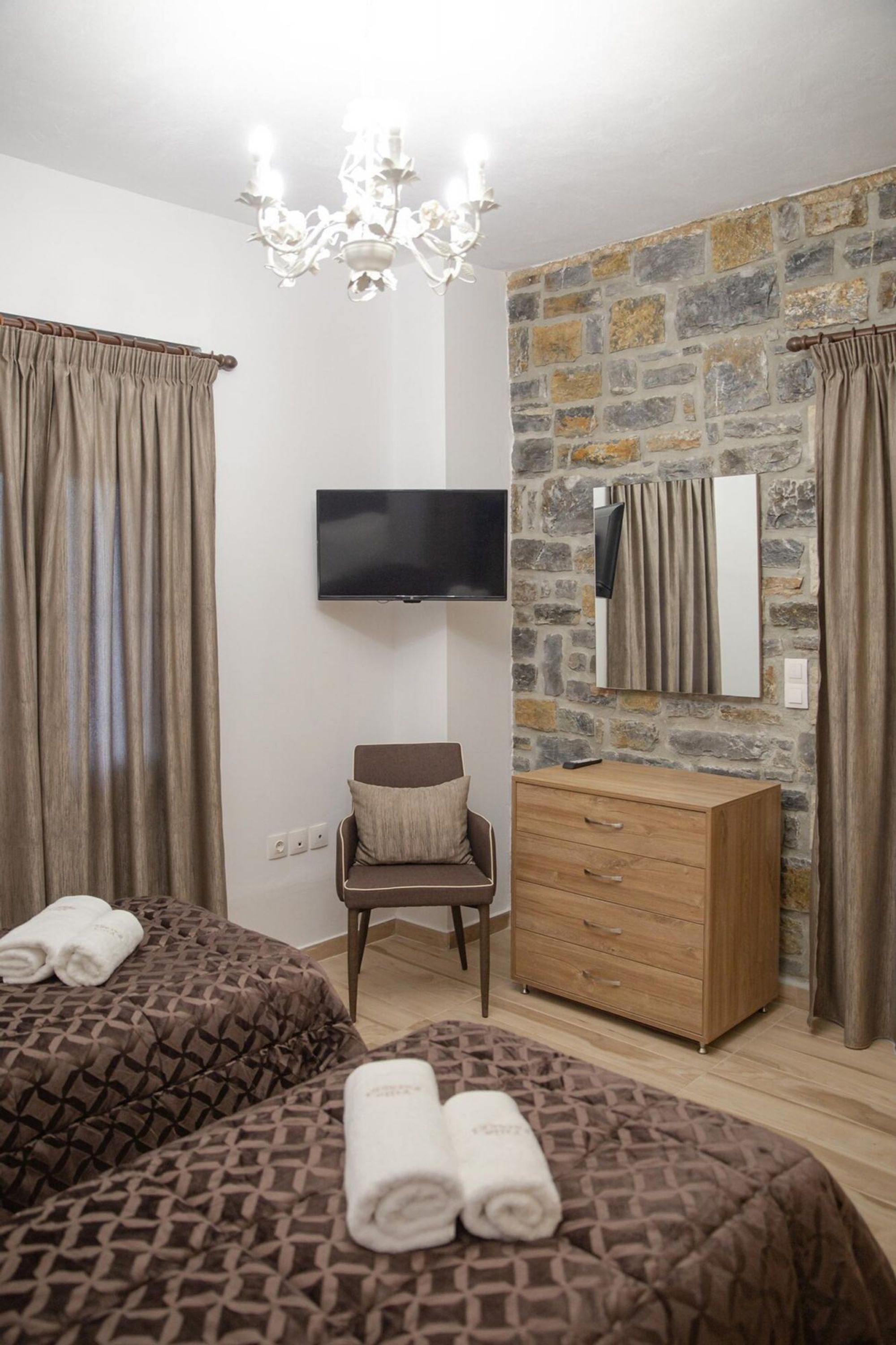 A hotel room with two beds, a TV, a wooden dresser, a chair, and a stone wall.
