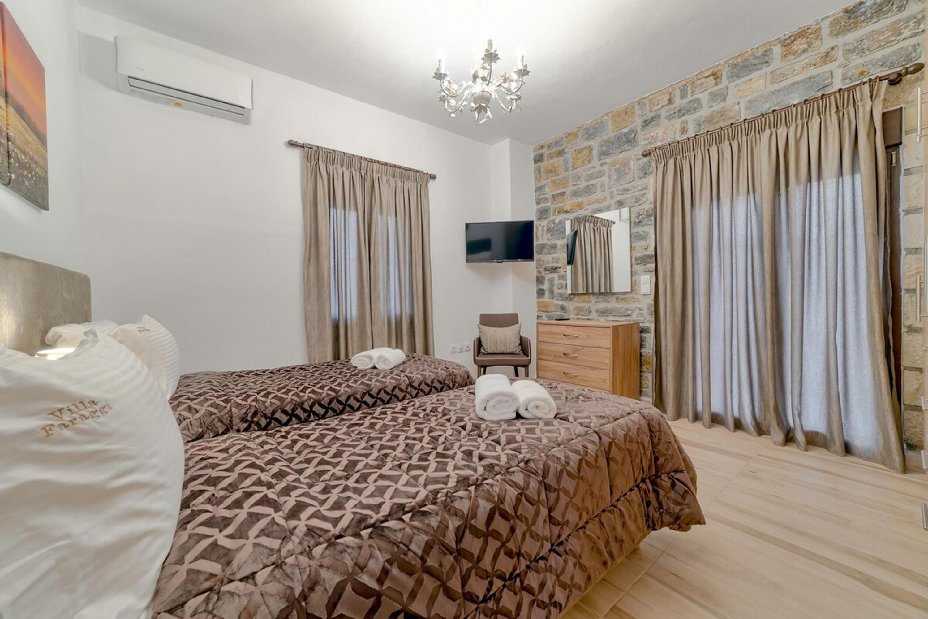 A hotel room with two beds, a TV, a stone wall, and a chandelier.
