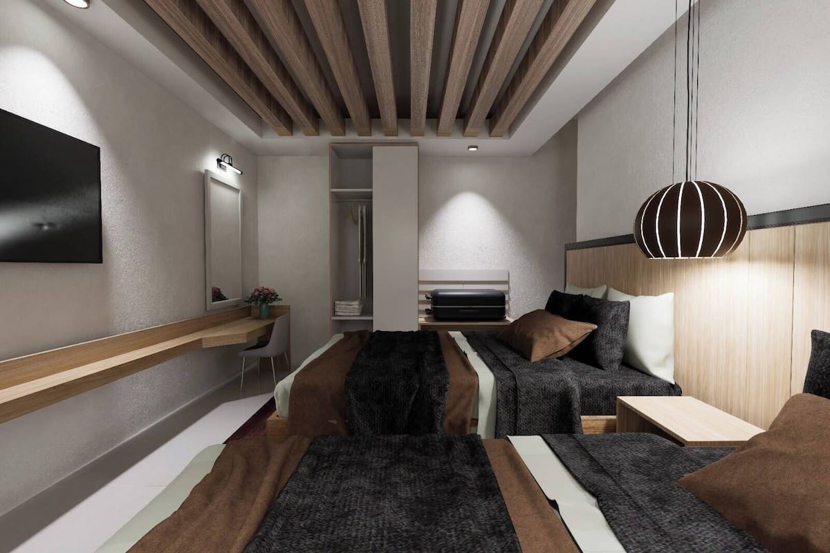A modern hotel room with a wooden ceiling, a flat-screen TV, a bed with multiple pillows, a bedside table, and a hanging light fixture.