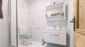 Un baño con cabina de ducha de vidrio, un inodoro con tapa azul, un lavamanos blanco con espejo encima y una puerta de madera.