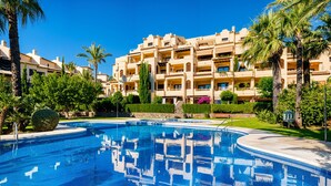 A swimming pool with a reflection of a multi-story building, surrounded by greenery and palm trees. - Apartment 'Villa Gadea 7-1' with Shared Pool, Private Terrace and Wi-Fi (Villa Gadea urbanizacion)