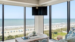 Habitació amb finestres grans amb vista a una platja, un televisor de pantalla plana fixat a la paret i una taula de centre de fusta amb una planta.